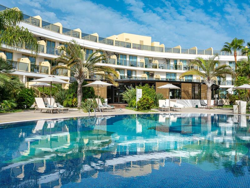 Modern hotel met groot zwembad en palmbomen onder een blauwe lucht.