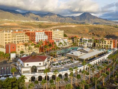 Duży hotel ośrodka z basenem i ogrodem palmowym w górskim krajobrazie pod pochmurnym niebem.