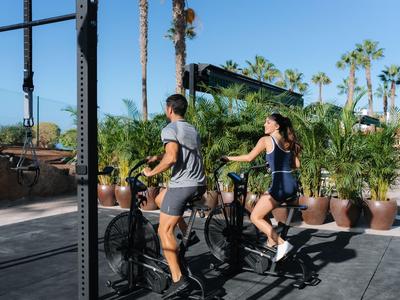 Due persone si allenano su cyclette all'aperto circondate da palme e cielo blu.