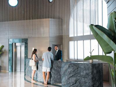 Reception dell'hotel con una coppia assistita da un membro dello staff, grandi finestre sullo sfondo.
