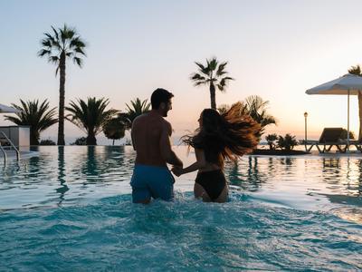 Coppia che balla in piscina al tramonto, circondata da palme e lettini.