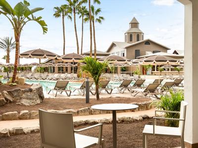 Vista di un'area piscina dell'hotel con lettini, ombrelloni e palme al sole.