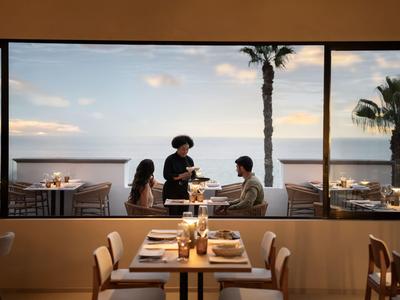 Vista attraverso una grande finestra su una terrazza con palme e ospiti, interno di un ristorante.