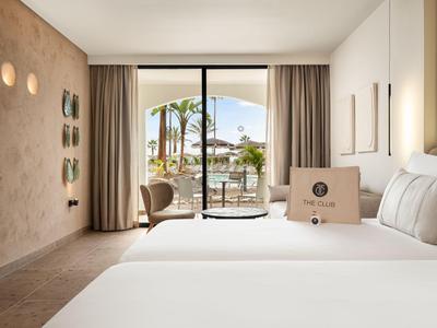 Camera d'hotel moderna con letti grandi, vista su una terrazza con palme attraverso una porta finestra scorrevole.