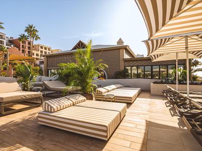 Terrazza di hotel soleggiata con lettini e ombrelloni vicino a palme e edifici.