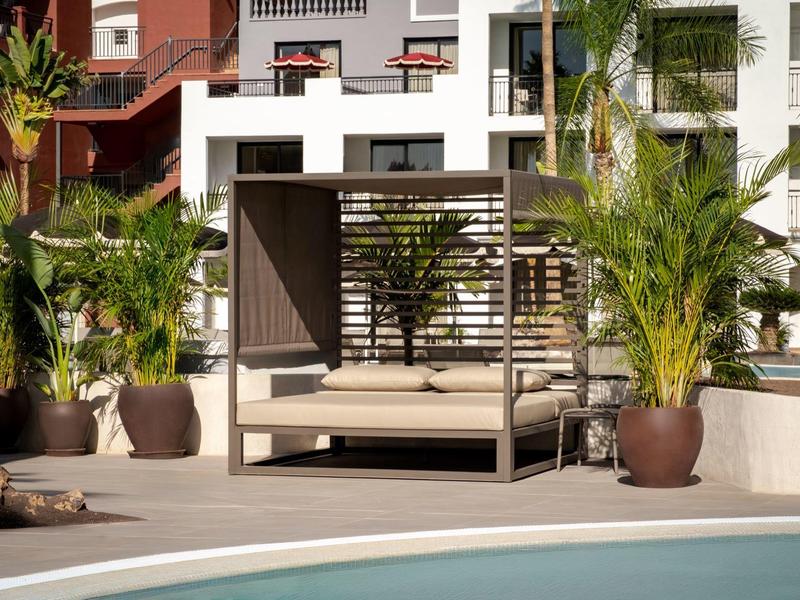 Nowoczesne łóżko na zewnątrz z osłoną przeciwsłoneczną i roślinami przy basenie hotelowym.