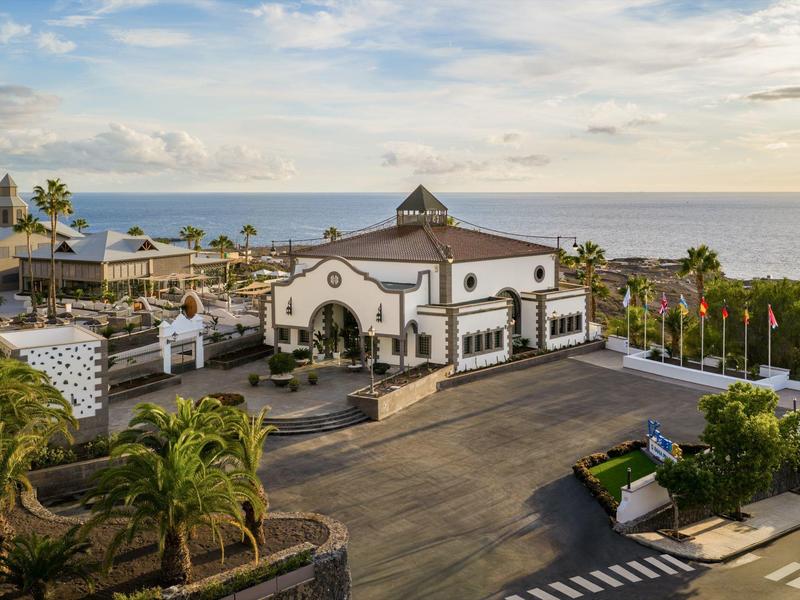 Budynek hotelu z białą elewacją i ciemnymi dachami nad morzem, otoczony palmami.