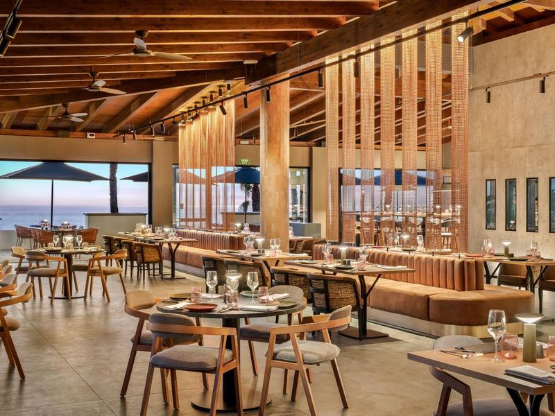 Nowoczesna restauracja z drewnianymi elementami, dużymi oknami i widokiem na morze o zachodzie słońca.