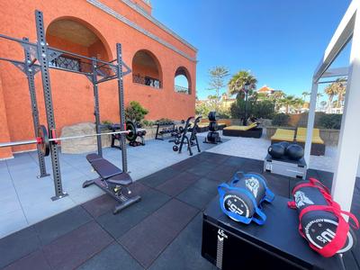 Nowoczesna siłownia na zewnątrz z urządzeniami i ciężarami przed pomarańczowym budynkiem.