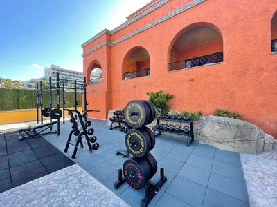 Nowoczesna siłownia na świeżym powietrzu z hantlami i sprzętem do ćwiczeń przed pomarańczowym budynkiem hotelu.