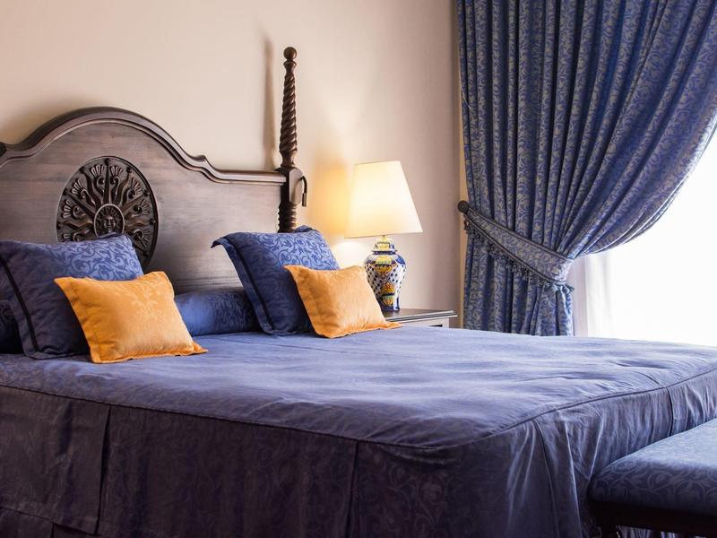 Łóżko podwójne z niebieską pościelą, dekoracyjnymi poduszkami i lampką nocną w pokoju z zasłonami.