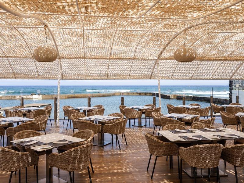 Otwarta restauracja z plecionymi krzesłami i stołami pod strzechą z widokiem na morze.