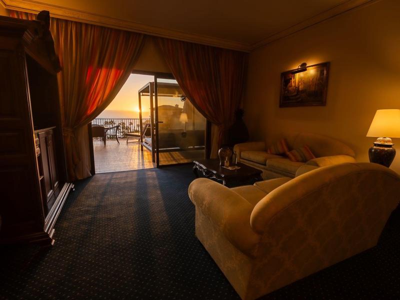 Przytulny pokój hotelowy z sofą, szafką na telewizor i tarasem z widokiem na morze o zachodzie słońca.