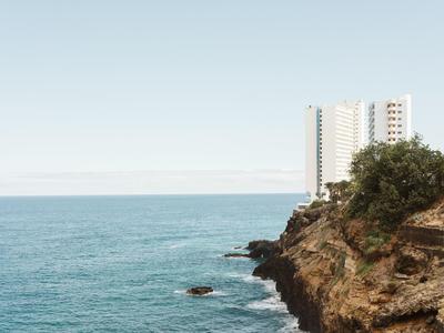 Costa rocciosa con un edificio alto sullo sfondo e mare calmo sotto un cielo azzurro.