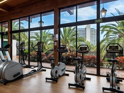 Salle de sport moderne avec vélos d'appartement et vue sur des palmiers et des bâtiments.