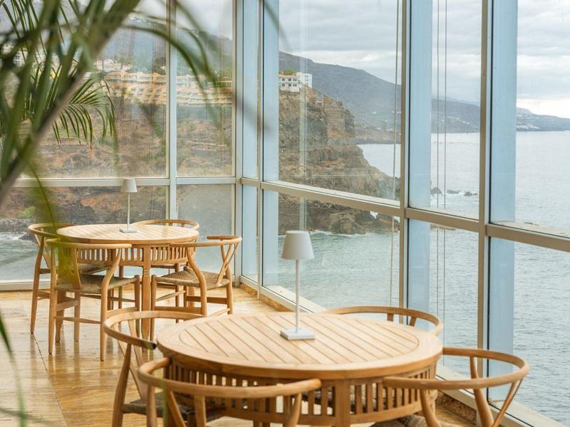 Area pranzo con tavoli e sedie in legno con vista sul mare attraverso grandi finestre di vetro.
