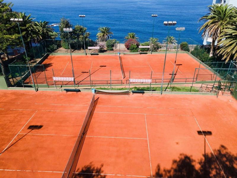 Più campi da tennis in terra rossa con palme e mare sullo sfondo.