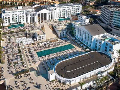 Vista aerea di un grande resort con piscine, lettini e edifici bianchi circondati da palme.