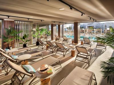 Lounge soleggiata accanto alla piscina dell'hotel con sedie moderne, piante di palma e area relax coperta.