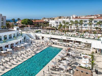 Groot hotel met buitenzwembad en veel ligstoelen onder een blauwe lucht.