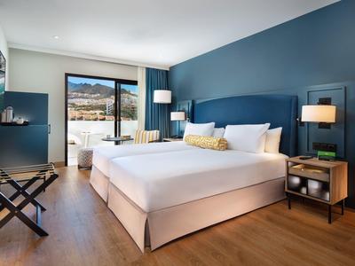 Camera d'hotel moderna con letto grande, parete blu, pavimento in legno e balcone con vista sulle montagne.