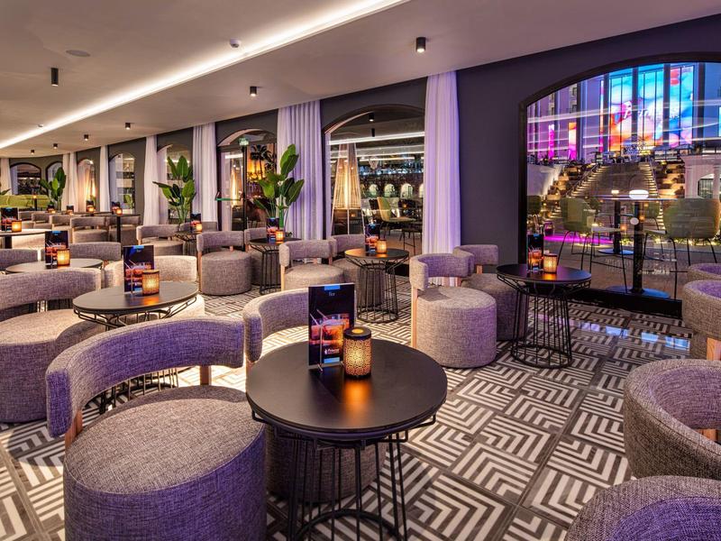 Lounge d'hotel moderno con poltrone viola, tappeto decorato e grandi finestre con luci colorate.