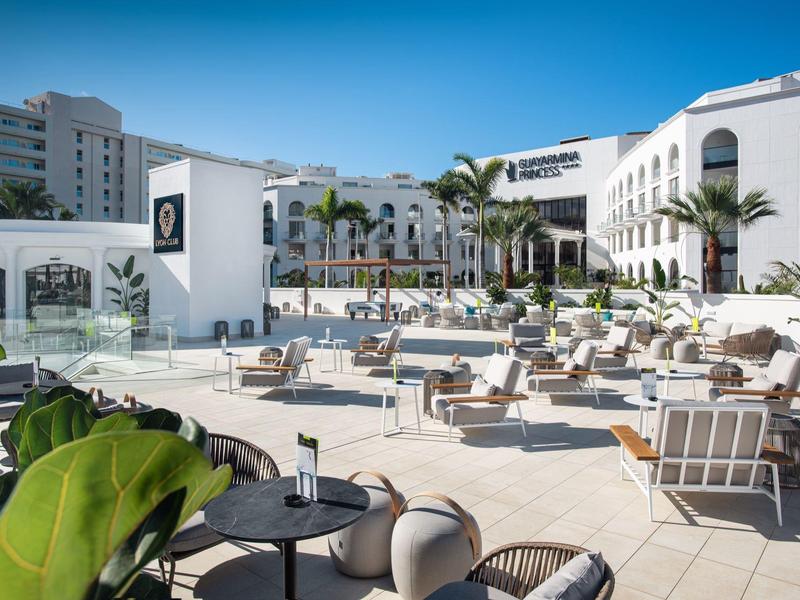 Ampia terrazza esterna dell'hotel con lettini, tavoli e palme sotto un cielo terso.