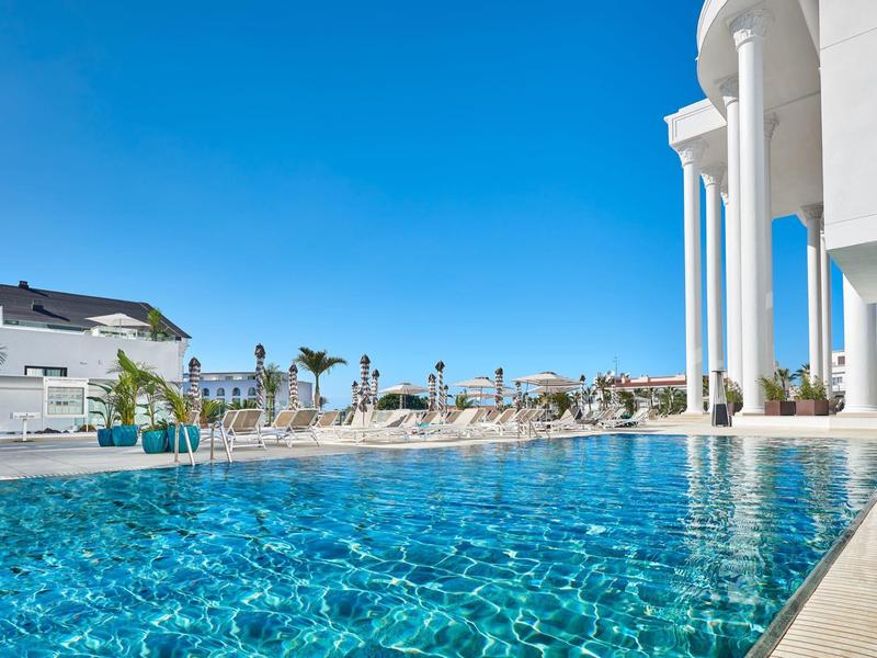 Piscina azzurra chiara accanto a un edificio dell'hotel bianco sotto un cielo soleggiato.