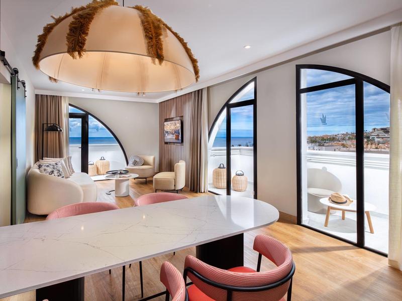 Suite d'hotel moderna con grandi finestre, vista oceano, area salotto e tavolo da pranzo con sedie rosa.