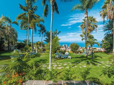 Jardín verde del hotel con palmeras, tumbonas y vista al mar azul bajo un cielo despejado.
