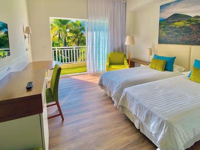 Habitación de hotel luminosa con dos camas, escritorio, sillón y acceso a balcón con vista a plantas.