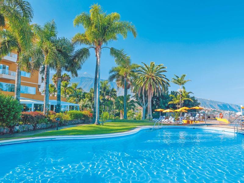 Piscina clara con palmeras y sombrillas amarillas junto a un hotel bajo cielo azul.