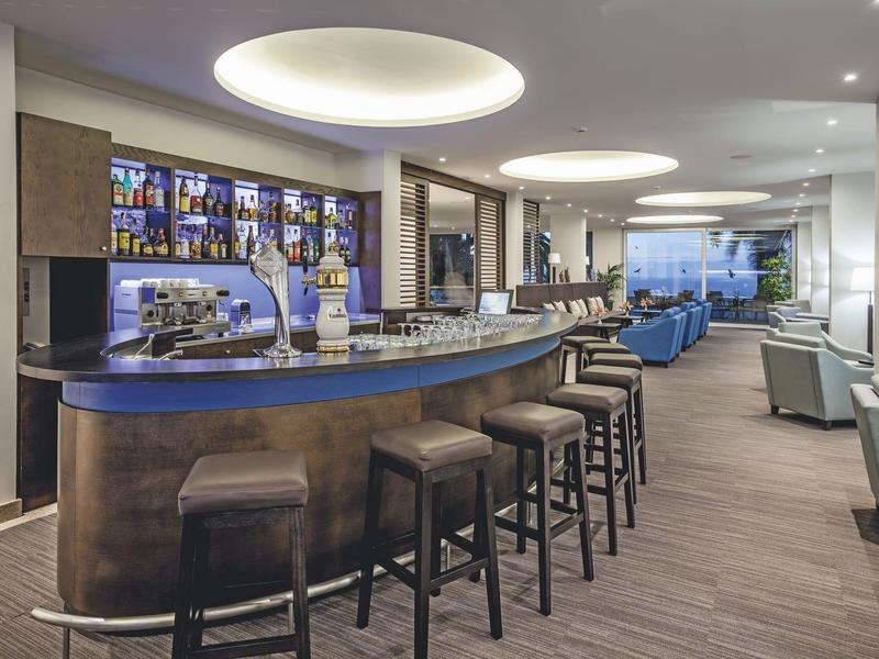 Área de bar moderna en un hotel con taburetes, barra iluminada y asientos al fondo.