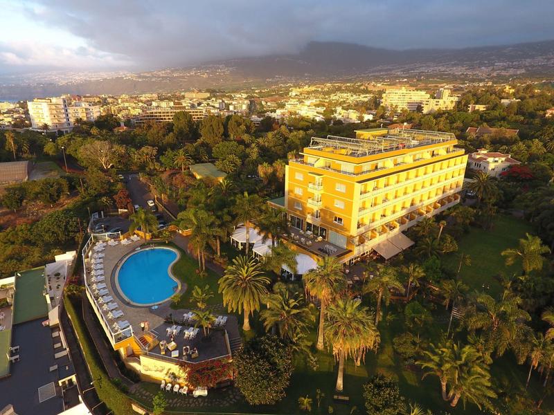 Hotel con edificio amarillo y piscina curva, rodeado de palmeras y paisaje urbano al fondo.