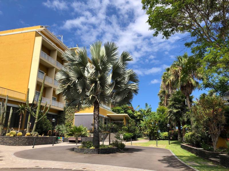 Edificio de hotel amarillo junto a jardín tropical con palmeras y cielo azul.