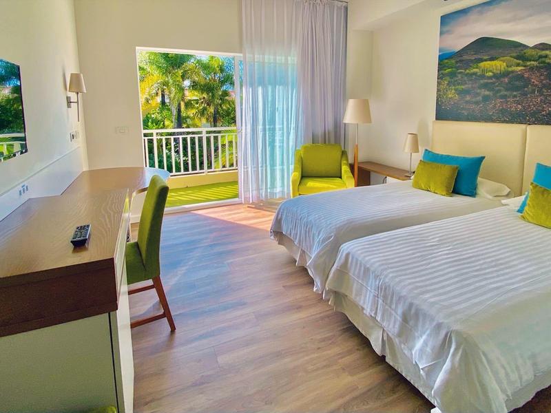 Habitación de hotel luminosa con dos camas, escritorio, sillón y acceso a balcón con vista a plantas.