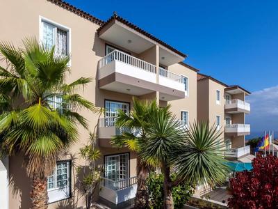 Vícepodlažní hotelová budova s balkóny, palmami a jasně modrou oblohou.