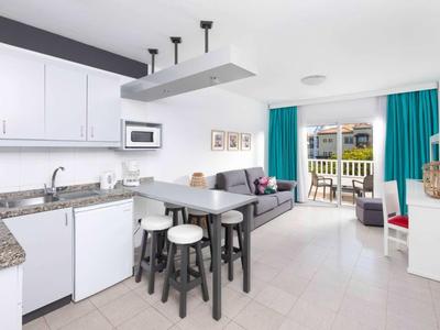 Světelná kuchyň hotelového pokoje s bílým nábytkem, barovými stoličkami a přístupem na balkon s výhledem na město.