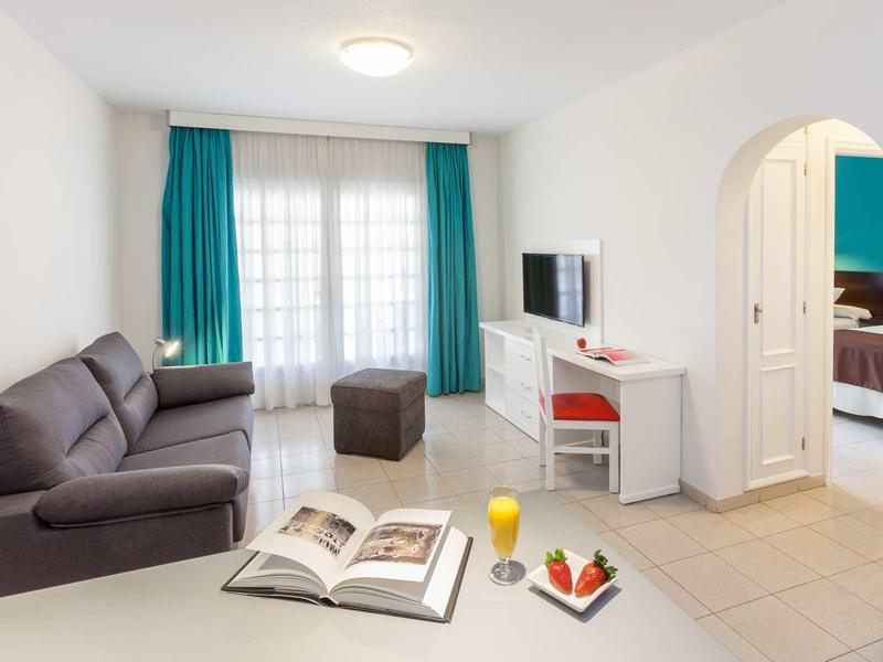 Moderně zařízený obývací pokoj s pohovkou, televizí, psacím stolem a přístupem do ložnice.