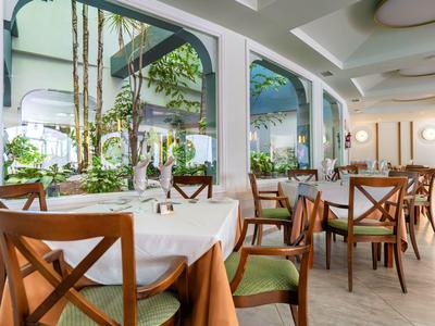 Salle à manger d'hôtel lumineuse avec chaises en bois, nappes blanches et grandes fenêtres avec vue sur verdure.