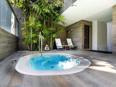 Jacuzzi intérieur avec eau bleue, murs en bois et deux chaises longues près des plantes.