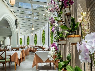 Véranda élégante d'hôtel avec plafond en verre, tables aux nappes beige et orchidées violettes.