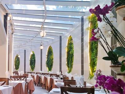 Salle à manger lumineuse avec tables en bois, nappes roses et hautes plantes vertes près des murs en verre.