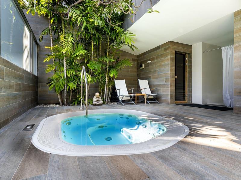 Jacuzzi intérieur avec eau bleue, murs en bois et deux chaises longues près des plantes.
