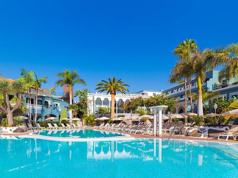 Piscine extérieure d'hôtel entourée de chaises longues et de palmiers sous un ciel bleu clair.