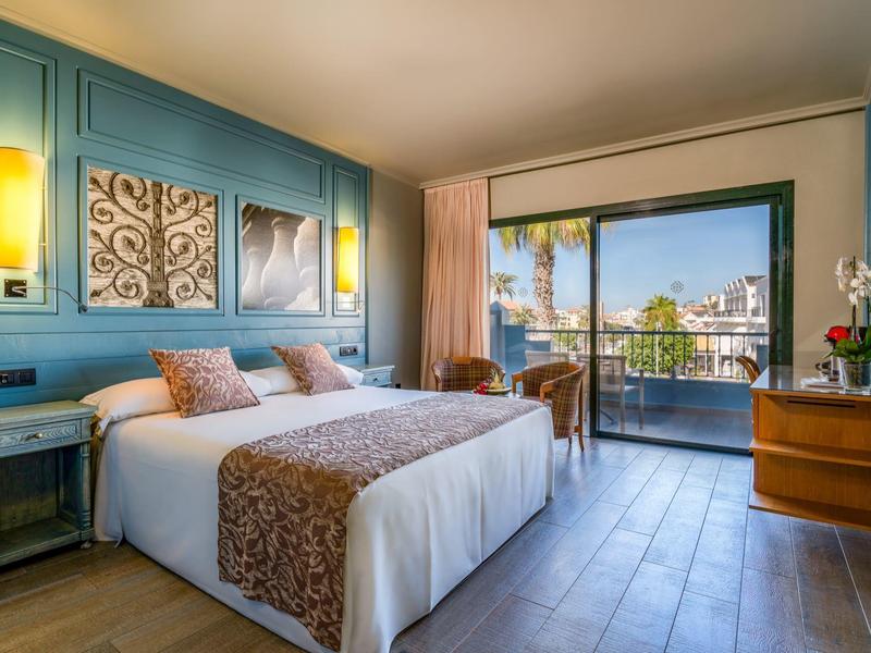 Chambre d'hôtel lumineuse avec lit double, murs bleus, sol en bois et balcon avec vue sur des palmiers.
