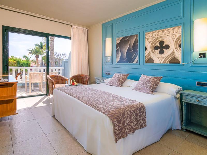 Chambre d'hôtel lumineuse avec grand lit, mur accent bleu, œuvres d'art et balcon avec vue sur les palmiers.