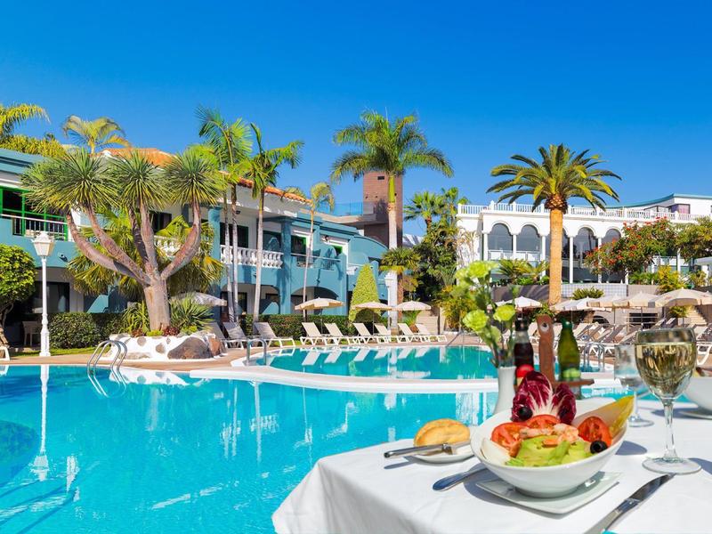 Piscine d'hôtel ensoleillée avec palmiers et table dressée avec boissons et fruits au premier plan.