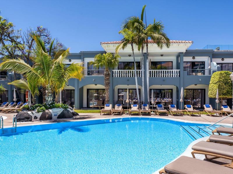 Hôtel ensoleillé avec piscine bleue, palmiers, chaises longues et ciel clair en arrière-plan.