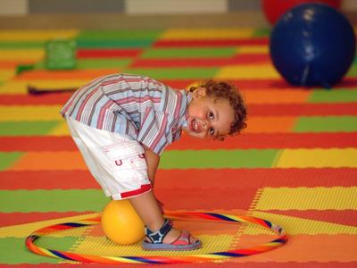 Ein Junge beugt sich in einem Hula-Hoop-Reifen auf bunter Spielfläche mit großen Bällen.